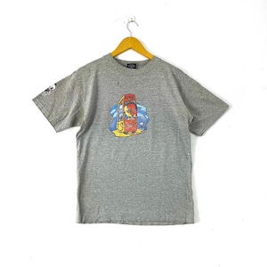 K&ouml;nnte beinhalten: Ein graues T-Shirt mit kurzen &Auml;rmeln und einem Grafikdruck eines roten und gelben Comic-Charakters in einer Telefonzelle. Der Charakter tr&auml;gt einen blauen Hut und eine Sonnenbrille. Die Grafik ist vor einem blauen und wei&szlig;en Hintergrund platziert.