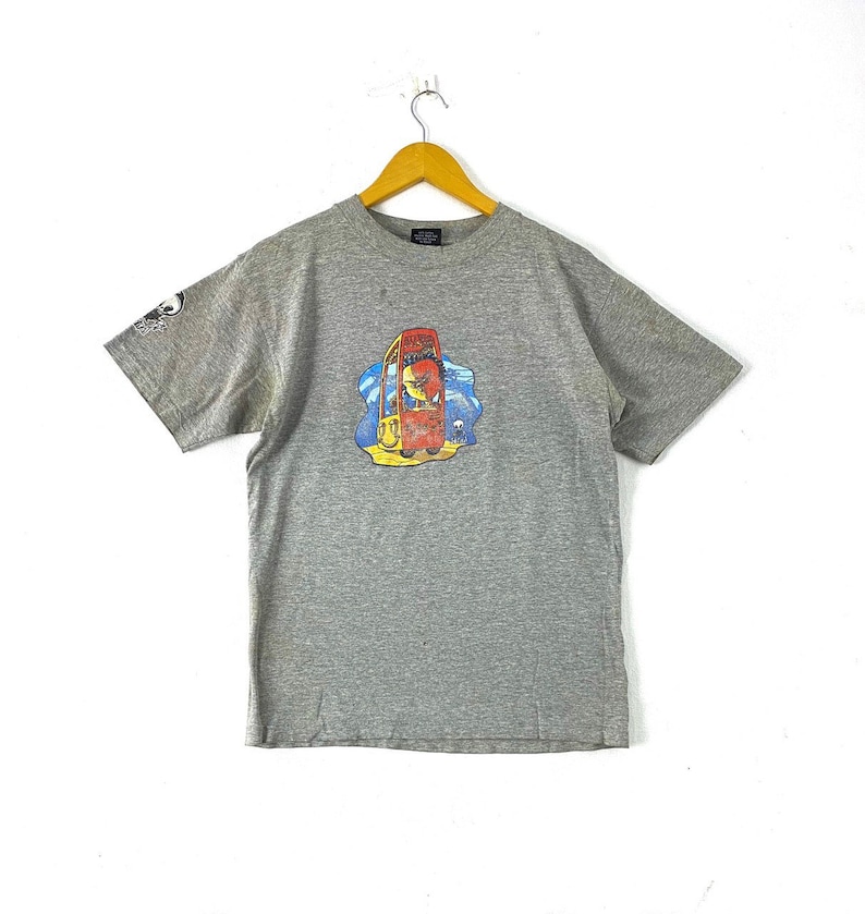 K&ouml;nnte beinhalten: Ein graues T-Shirt mit kurzen &Auml;rmeln und einem Grafikdruck eines roten und gelben Comic-Charakters in einer Telefonzelle. Der Charakter tr&auml;gt einen blauen Hut und eine Sonnenbrille. Die Grafik ist vor einem blauen und wei&szlig;en Hintergrund platziert.