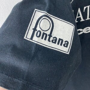 Puede incluir: Camiseta negra con un gr&aacute;fico blanco de un c&iacute;rculo con una l&iacute;nea que lo atraviesa y la palabra "fontana" debajo. El texto "The Great Race" es visible en el fondo.