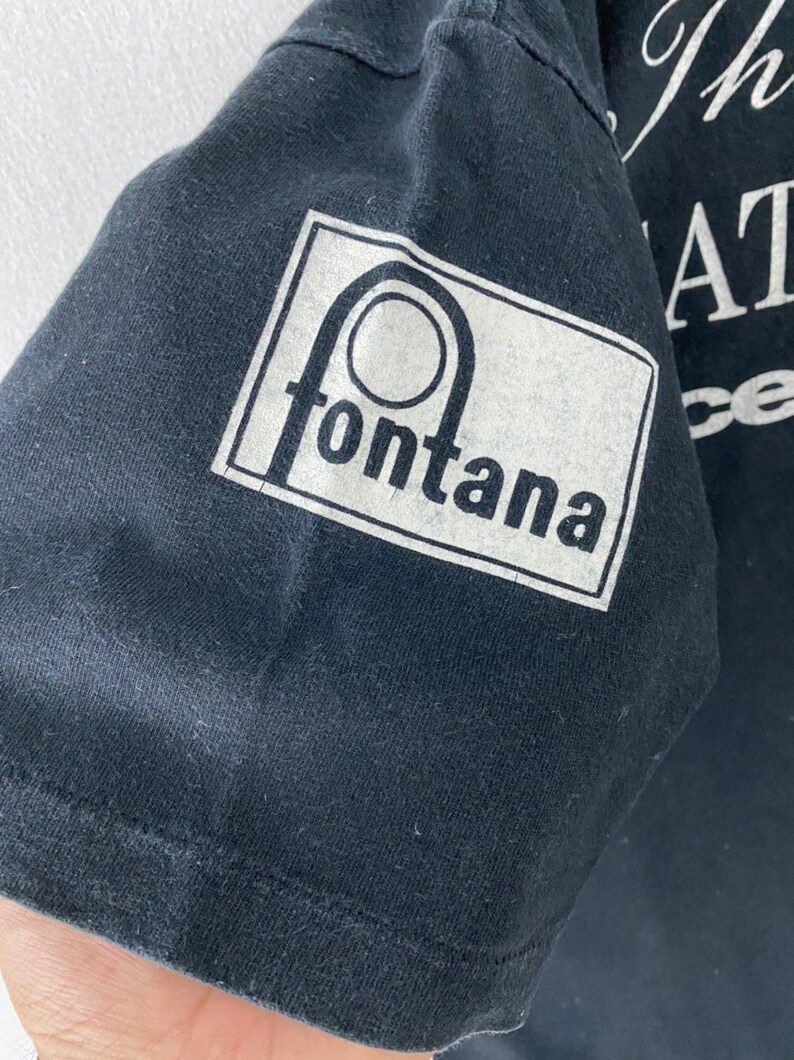 Puede incluir: Camiseta negra con un gr&aacute;fico blanco de un c&iacute;rculo con una l&iacute;nea que lo atraviesa y la palabra "fontana" debajo. El texto "The Great Race" es visible en el fondo.