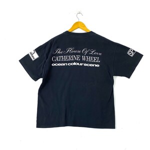 Puede incluir: Camiseta negra con texto blanco que dice "The House of Love Catherine Wheel ocean colourscene". La camiseta tiene un logotipo blanco en las mangas izquierda y derecha que dice "S" en un c&iacute;rculo.