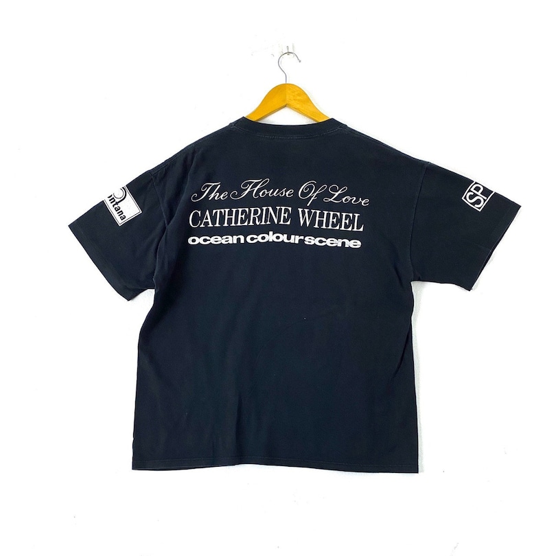 Puede incluir: Camiseta negra con texto blanco que dice "The House of Love Catherine Wheel ocean colourscene". La camiseta tiene un logotipo blanco en las mangas izquierda y derecha que dice "S" en un c&iacute;rculo.