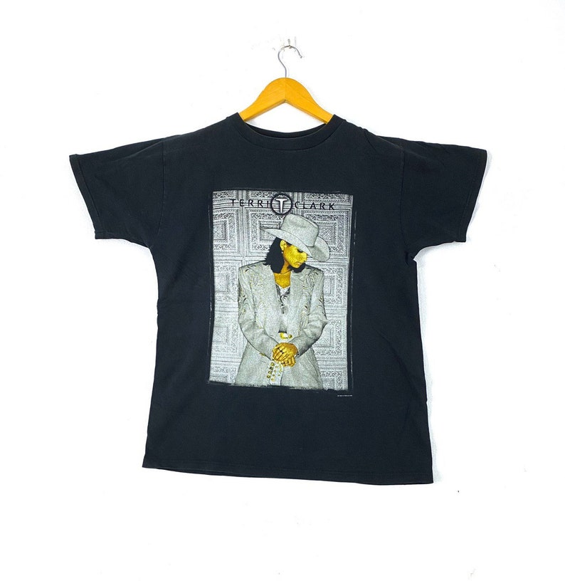 Może przedstawiać: Czarny T-shirt z grafiką piosenkarki Terri Clark w białym kapeluszu i srebrnej kurtce. Tekst "Terri Clark" jest nadrukowany na grafice złotem.
