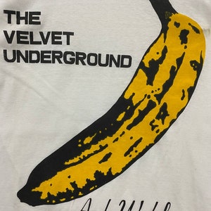 May include: White t-shirt featuring a black and yellow banana graphic with the text "THE VELVET UNDERGROUND" and "Andy Warhol" printed on it.