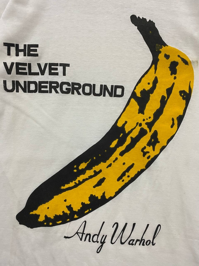 May include: White t-shirt featuring a black and yellow banana graphic with the text "THE VELVET UNDERGROUND" and "Andy Warhol" printed on it.
