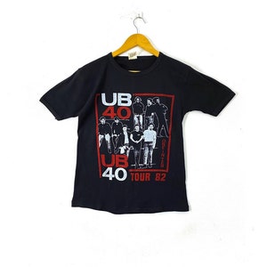 Puede incluir: Camiseta negra con un estampado gráfico blanco y rojo de la banda UB40. El gráfico incluye una foto de los miembros de la banda y el texto "UB40 One In Ten Tour 82".