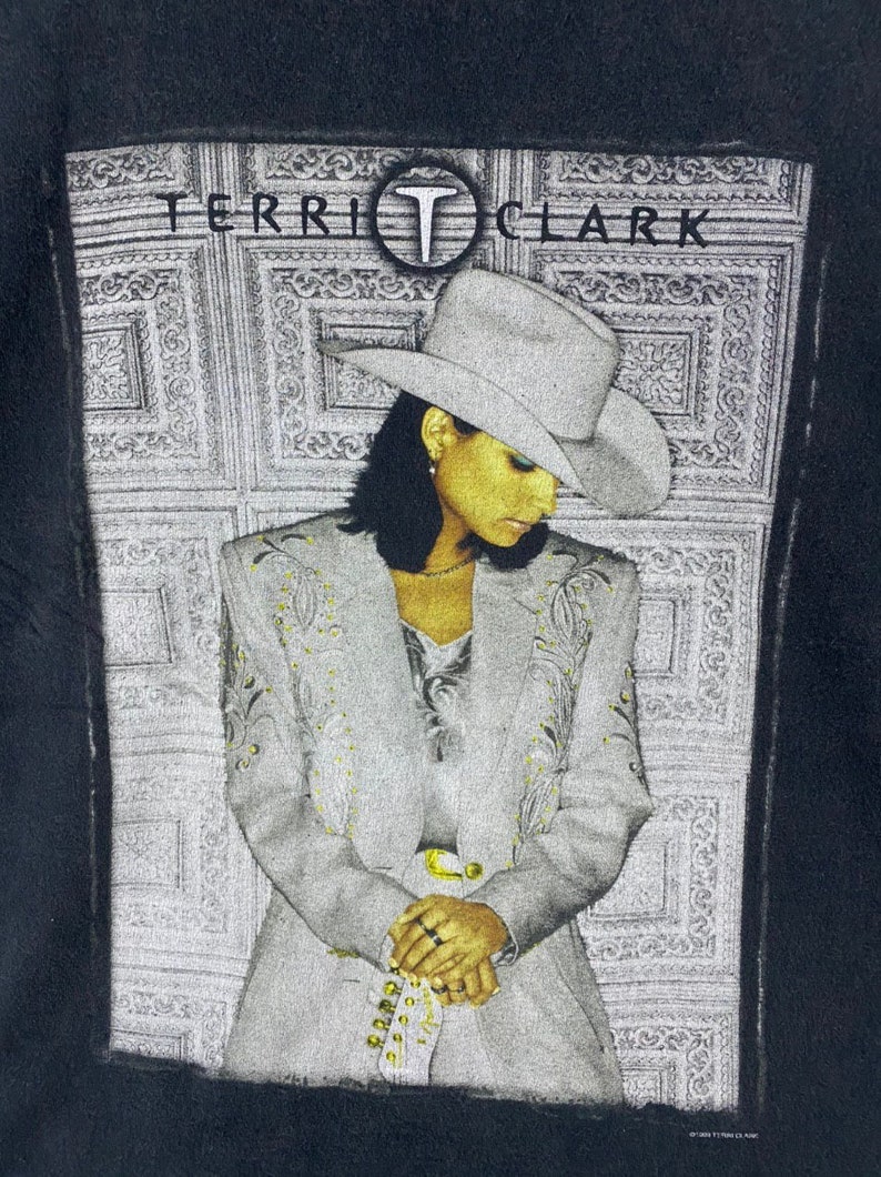 Może przedstawiać: Czarny T-shirt z białym obrazem kobiety w białym kowbojskim kapeluszu i srebrnej kurtce. Tekst "TERRI CLARK" jest nadrukowany na g&oacute;rze obrazu czarnym kolorem.