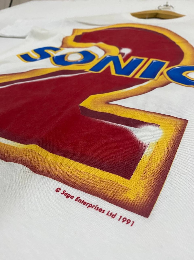 May include: White t-shirt with a red and yellow graphic of the number "2" with the word "SONIC" in blue letters above it. The text "&copy; Sega Enterprises Ltd 1991" is printed below the graphic.