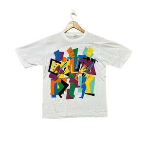 Op de afbeelding: Wit T-shirt met een kleurrijk abstract ontwerp van vier figuren die muziekinstrumenten bespelen. Het ontwerp bevat geometrische vormen in tinten geel, oranje, rood, paars, blauw, groen en zwart.