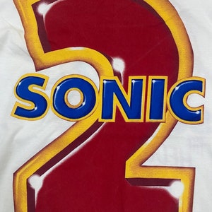 May include: A white t-shirt with a large red number "2" outlined in yellow. The word "SONIC" is written in blue letters across the number. The text "Sega Enterprises Ltd 1991" is at the bottom of the shirt.