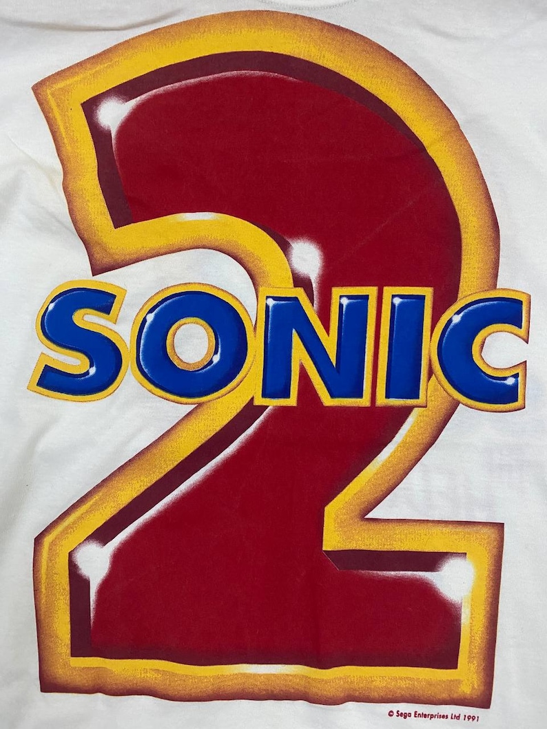 May include: A white t-shirt with a large red number "2" outlined in yellow. The word "SONIC" is written in blue letters across the number. The text "Sega Enterprises Ltd 1991" is at the bottom of the shirt.