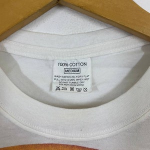May include: White t-shirt with a red, yellow, and orange graphic design. The tag says "100% Cotton, Medium, Wash Separately/Dry Flat, Pull into Shape When Wet, Do Not Tumble Dry, Do Not Iron Motif."