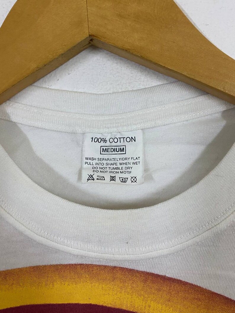 May include: White t-shirt with a red, yellow, and orange graphic design. The tag says "100% Cotton, Medium, Wash Separately/Dry Flat, Pull into Shape When Wet, Do Not Tumble Dry, Do Not Iron Motif."