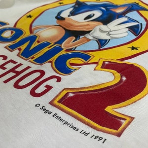 May include: White t-shirt with a yellow circle and a blue cartoon character of Sonic the Hedgehog. The text "Sonic the Hedgehog 2" is printed in red and yellow. The copyright text "&copy; Sega Enterprises Ltd 1991" is printed below the circle.