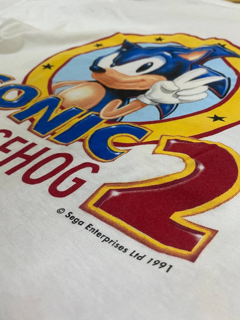 May include: White t-shirt with a yellow circle and a blue cartoon character of Sonic the Hedgehog. The text "Sonic the Hedgehog 2" is printed in red and yellow. The copyright text "&copy; Sega Enterprises Ltd 1991" is printed below the circle.