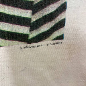Puede incluir: Una camiseta blanca con un dise&ntilde;o de rayas blancas y negras. La camiseta tiene el texto "&copy;1985 Areagraph Ltd. Pet Shop Boys" impreso en la parte inferior.