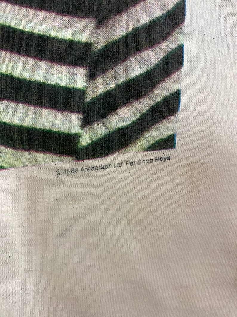 Puede incluir: Una camiseta blanca con un dise&ntilde;o de rayas blancas y negras. La camiseta tiene el texto "&copy;1985 Areagraph Ltd. Pet Shop Boys" impreso en la parte inferior.