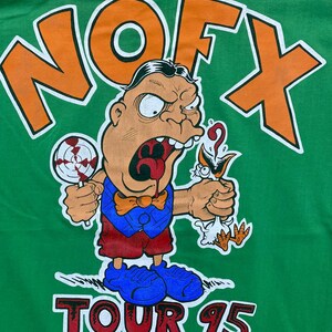 Puede incluir: Camiseta verde con un logotipo "NOFX" naranja y blanco y un personaje de dibujos animados que sostiene una piruleta y un p&aacute;jaro. El texto "TOUR 95" est&aacute; impreso debajo del personaje. El texto de copyright "&copy; 1995 NOFX UNDER LICENSE TO TRASH MARK" est&aacute; impreso en la parte inferior.