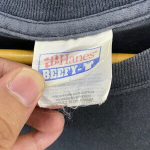 K&ouml;nnte beinhalten: Ein wei&szlig;es Hanes-Etikett mit blauem Text, der "Hanes" und "Beefy-T" in Fettdruck lautet. Das Etikett enth&auml;lt auch den Text "Preshrunk 100% Cotton Assembled in Mexico" in kleinerer Schrift.