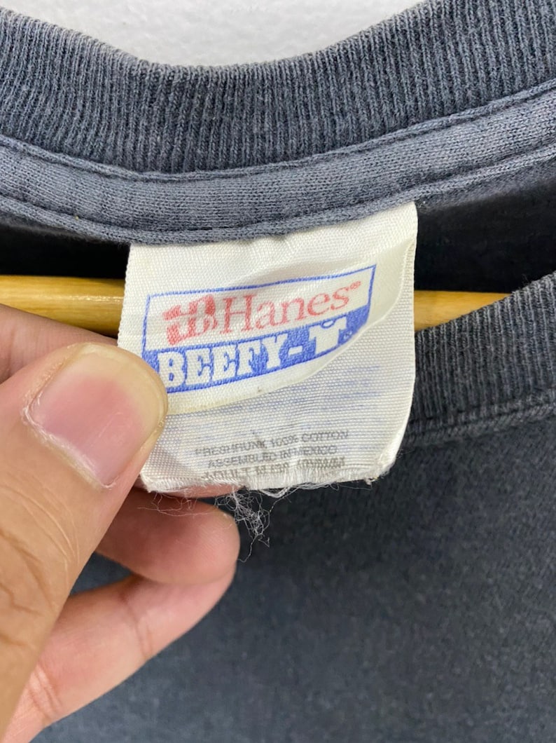 K&ouml;nnte beinhalten: Ein wei&szlig;es Hanes-Etikett mit blauem Text, der "Hanes" und "Beefy-T" in Fettdruck lautet. Das Etikett enth&auml;lt auch den Text "Preshrunk 100% Cotton Assembled in Mexico" in kleinerer Schrift.