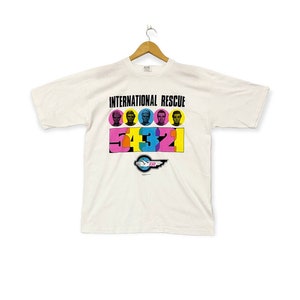 May include: White t-shirt with a graphic print of the International Rescue logo and the text "INTERNATIONAL RESCUE" in black. The logo features a white hand with a pink wing and the text "IR" in pink. The graphic also includes five colorful circles with the faces of the characters from the show, and the numbers 54321 in colorful blocks.