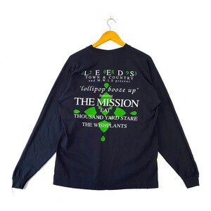 Peut inclure: T-shirt à manches longues noire avec un imprimé graphique vert comportant le texte "2L2 E 0E8 D 9S3 TOWN & COUNTRY and M.W.I.S present 'lollipop booze up' featuring THE MISSION EAT THOUSAND YARD STARE THE WISHPLANTS"