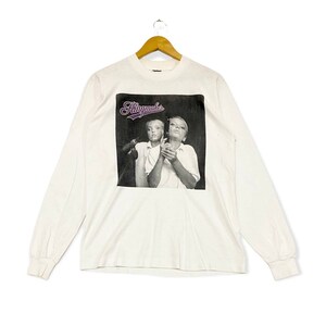 Può includere: Maglietta bianca a maniche lunghe con una foto in bianco e nero di due persone e il testo "Kingmaker" in viola.