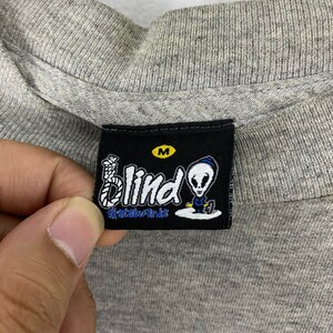 K&ouml;nnte beinhalten: Ein schwarz-wei&szlig;es Etikett mit dem Text "Blind Skateboards" und einem Comic-Alien mit einer Schlinge um den Hals. Das Etikett hat ein wei&szlig;es "M" in einem schwarzen Kreis.