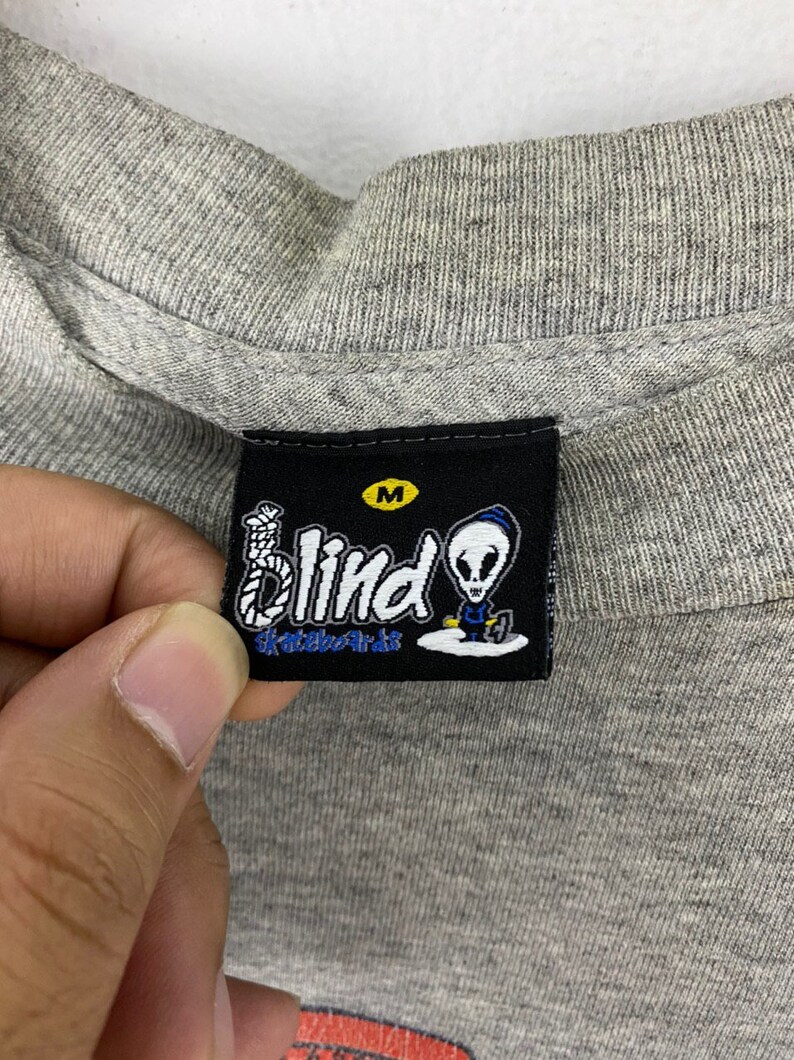 K&ouml;nnte beinhalten: Ein schwarz-wei&szlig;es Etikett mit dem Text "Blind Skateboards" und einem Comic-Alien mit einer Schlinge um den Hals. Das Etikett hat ein wei&szlig;es "M" in einem schwarzen Kreis.