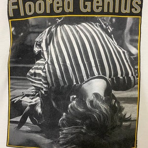Pu&ograve; includere: Foto in bianco e nero di una persona sdraiata sulla schiena con la testa rivolta verso la fotocamera. La persona indossa una camicia a righe e ha i capelli che le coprono il viso. La foto &egrave; incorniciata da un bordo nero e giallo con il testo "Floored Genius" sopra la foto.