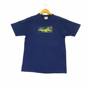 May include: A navy blue t-shirt with a graphic print of a car driving through a drive-in movie theater. The text "at the drive-in" is printed in green and blue.