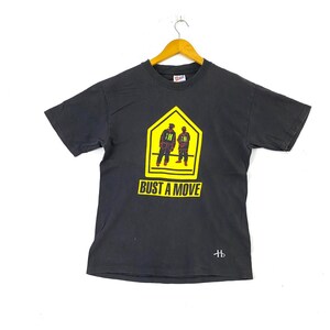 May include: Black t-shirt with a yellow and black warning sign graphic featuring two figures in red and black clothing. The text "Bust a Move" is printed below the sign.