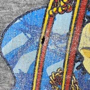 K&ouml;nnte beinhalten: Nahaufnahme eines grauen T-Shirts mit einem verblassten Grafikdruck eines Comic-Charakters, der ein Surfbrett h&auml;lt. Der Charakter tr&auml;gt ein rot-gelbes Shirt und eine blaue Hose. Der Hintergrund ist ein blau-wei&szlig;es Muster.