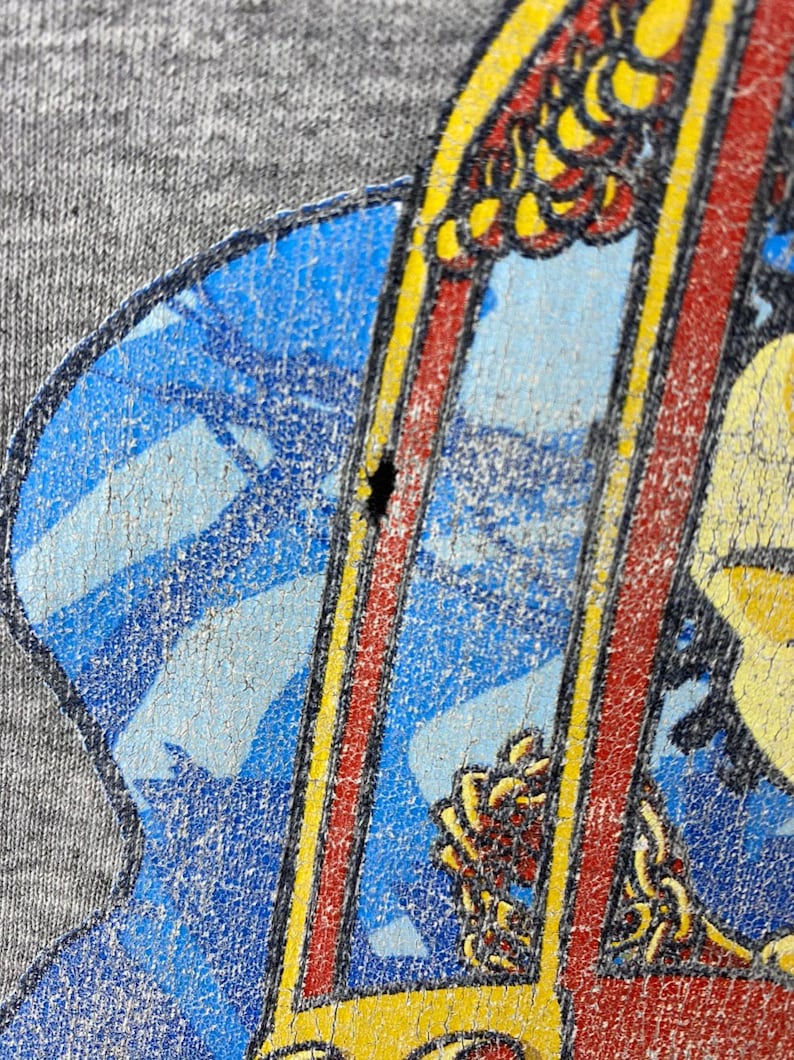 K&ouml;nnte beinhalten: Nahaufnahme eines grauen T-Shirts mit einem verblassten Grafikdruck eines Comic-Charakters, der ein Surfbrett h&auml;lt. Der Charakter tr&auml;gt ein rot-gelbes Shirt und eine blaue Hose. Der Hintergrund ist ein blau-wei&szlig;es Muster.