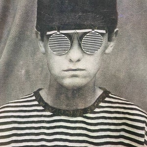 Puede incluir: Una foto en blanco y negro de una persona que lleva una camisa a rayas y una gorra negra con gafas de sol. Las gafas de sol tienen un dise&ntilde;o &uacute;nico con l&iacute;neas horizontales. El texto "POST ABOUT" es visible en la camisa.