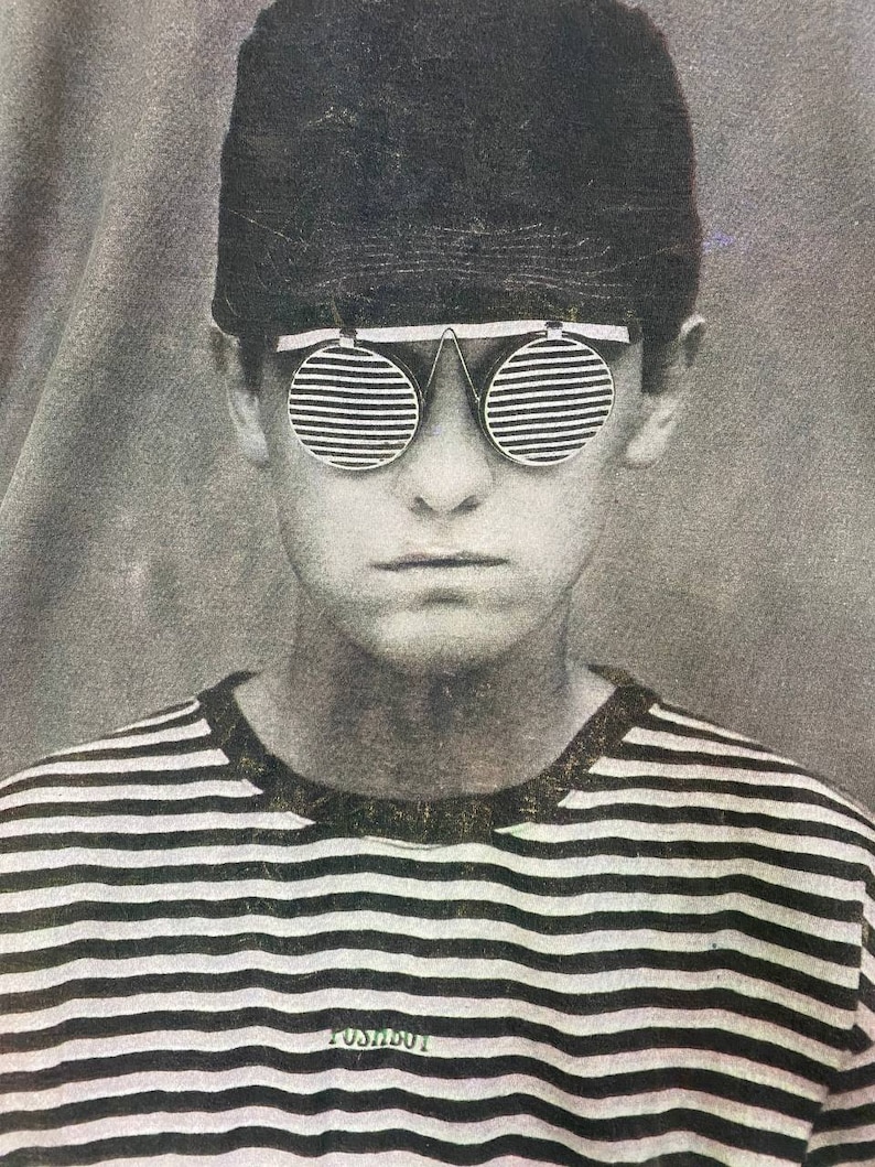 Puede incluir: Una foto en blanco y negro de una persona que lleva una camisa a rayas y una gorra negra con gafas de sol. Las gafas de sol tienen un dise&ntilde;o &uacute;nico con l&iacute;neas horizontales. El texto "POST ABOUT" es visible en la camisa.