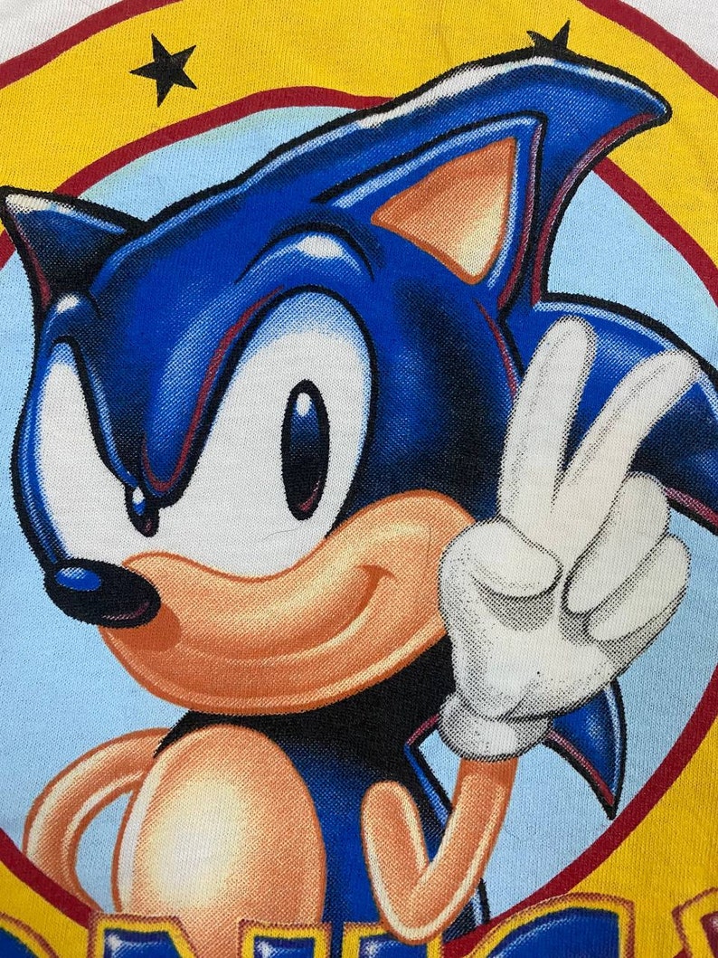 May include: A blue and yellow cartoon character with a red and yellow background. The character is Sonic the Hedgehog, and he is giving a peace sign with his right hand. The character is smiling and has a white glove on his right hand.