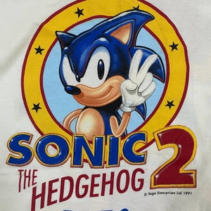 May include: A white t-shirt with a blue and red graphic of Sonic the Hedgehog from the video game Sonic the Hedgehog 2. The graphic features Sonic giving a peace sign with a yellow circle and stars around him. The text "Sonic the Hedgehog 2" is in blue and red, and "SEGA" is in blue. The copyright text "&copy; Sega Enterprises Ltd 1991" is in black.
