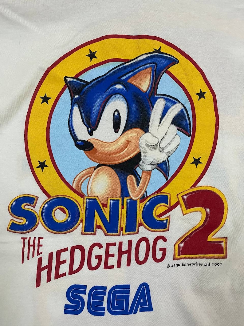 May include: A white t-shirt with a blue and red graphic of Sonic the Hedgehog from the video game Sonic the Hedgehog 2. The graphic features Sonic giving a peace sign with a yellow circle and stars around him. The text "Sonic the Hedgehog 2" is in blue and red, and "SEGA" is in blue. The copyright text "&copy; Sega Enterprises Ltd 1991" is in black.