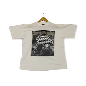 Pu&ograve; includere: Maglietta bianca con una foto in bianco e nero di una persona con una camicia a righe. Il testo "Floored Genius" &egrave; stampato in oro sopra la foto.