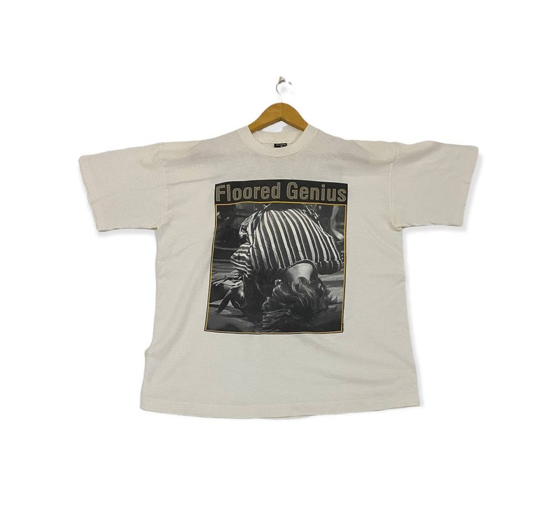 Pu&ograve; includere: Maglietta bianca con una foto in bianco e nero di una persona con una camicia a righe. Il testo "Floored Genius" &egrave; stampato in oro sopra la foto.