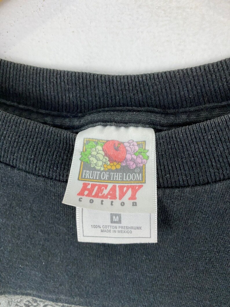 Może przedstawiać: Czarna metka koszulki Fruit of the Loom z białym tekstem, kt&oacute;ry brzmi "Heavy Cotton" i "100% Cotton Preshrunk Made in Mexico".