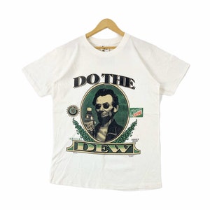Pode incluir: Camiseta branca com um gráfico verde e preto de Abraham Lincoln usando óculos de sol e segurando uma garrafa de Mountain Dew. O texto "Do the Dew" está acima do gráfico e "Series 2000" está abaixo.