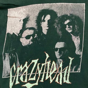 Puede incluir: Una camiseta negra con una imagen descolorida en blanco y gris de una banda llamada "Crazyhead". Los miembros de la banda llevan gafas de sol y tienen el pelo largo.
