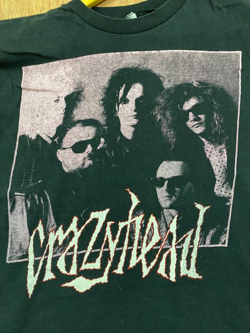 Puede incluir: Una camiseta negra con una imagen descolorida en blanco y gris de una banda llamada "Crazyhead". Los miembros de la banda llevan gafas de sol y tienen el pelo largo.