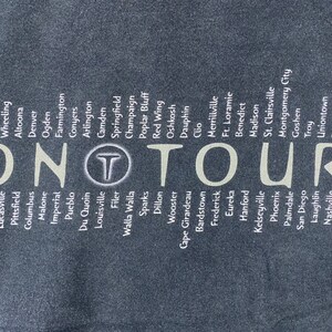 Może przedstawiać: Czarny T-shirt z białym tekstem wymieniającym miasta w Stanach Zjednoczonych. Tekst "ON TOUR" jest wyśrodkowany na koszulce.