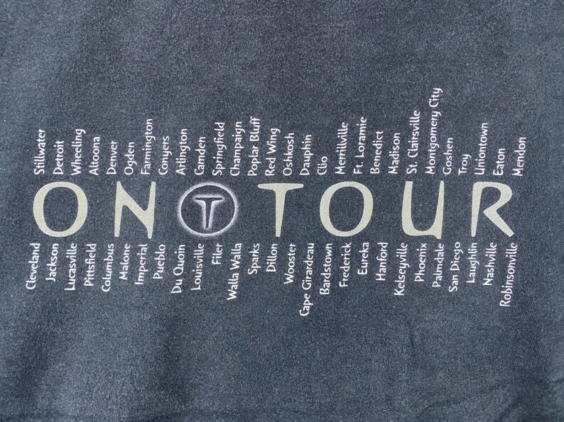 Może przedstawiać: Czarny T-shirt z białym tekstem wymieniającym miasta w Stanach Zjednoczonych. Tekst "ON TOUR" jest wyśrodkowany na koszulce.