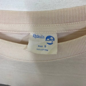 Puede incluir: Una camiseta rosa claro con una etiqueta azul y blanca que dice "Splash" y "Talla S 100% Algod&oacute;n".