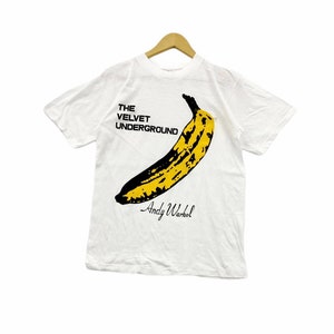 May include: White t-shirt featuring a yellow banana graphic with the text "The Velvet Underground" and "Andy Warhol" printed in black.