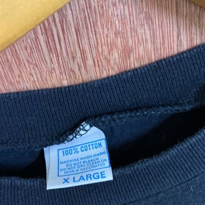 Puede incluir: Una camiseta negra con una etiqueta de cuidado azul que dice "100% Algod&oacute;n, Lavar a m&aacute;quina en caliente, No usar lej&iacute;a, No limpiar en seco o planchar si est&aacute; decorado, Talla XL."
